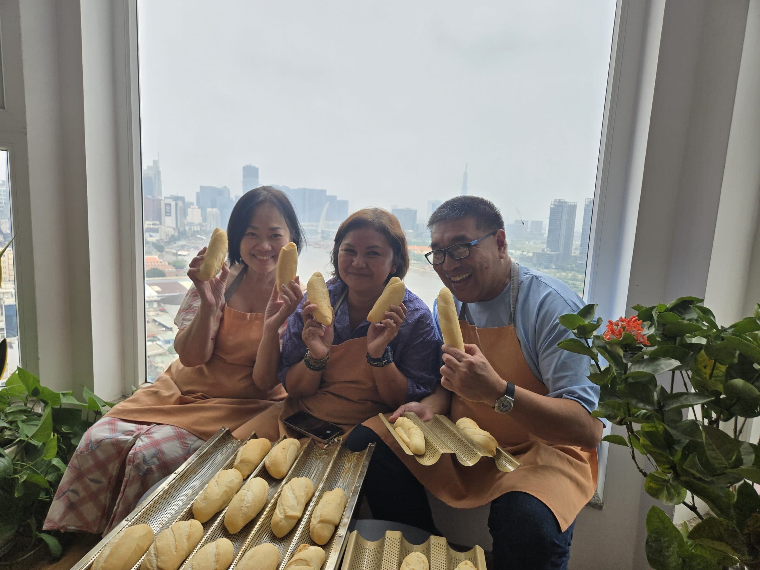 Three students hold finished bánh mì loaves at Lua's Kitchen baking class, with the Ho Chi Minh City skyline behind them