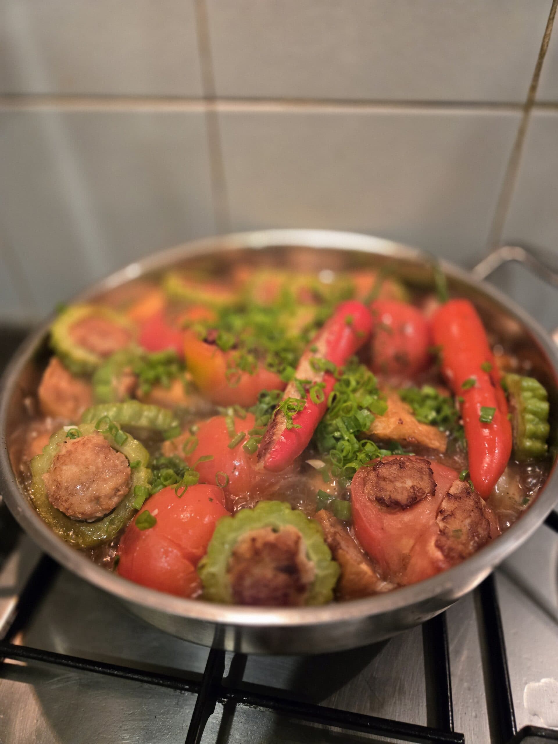 Yong Tau Foo simmering in a stainless pan on a gas hob: stuffed chillies, bitter gourd rings showing the seared pork-and-fish paste, stuffed tomatoes, and scattered spring onion in a clear stock