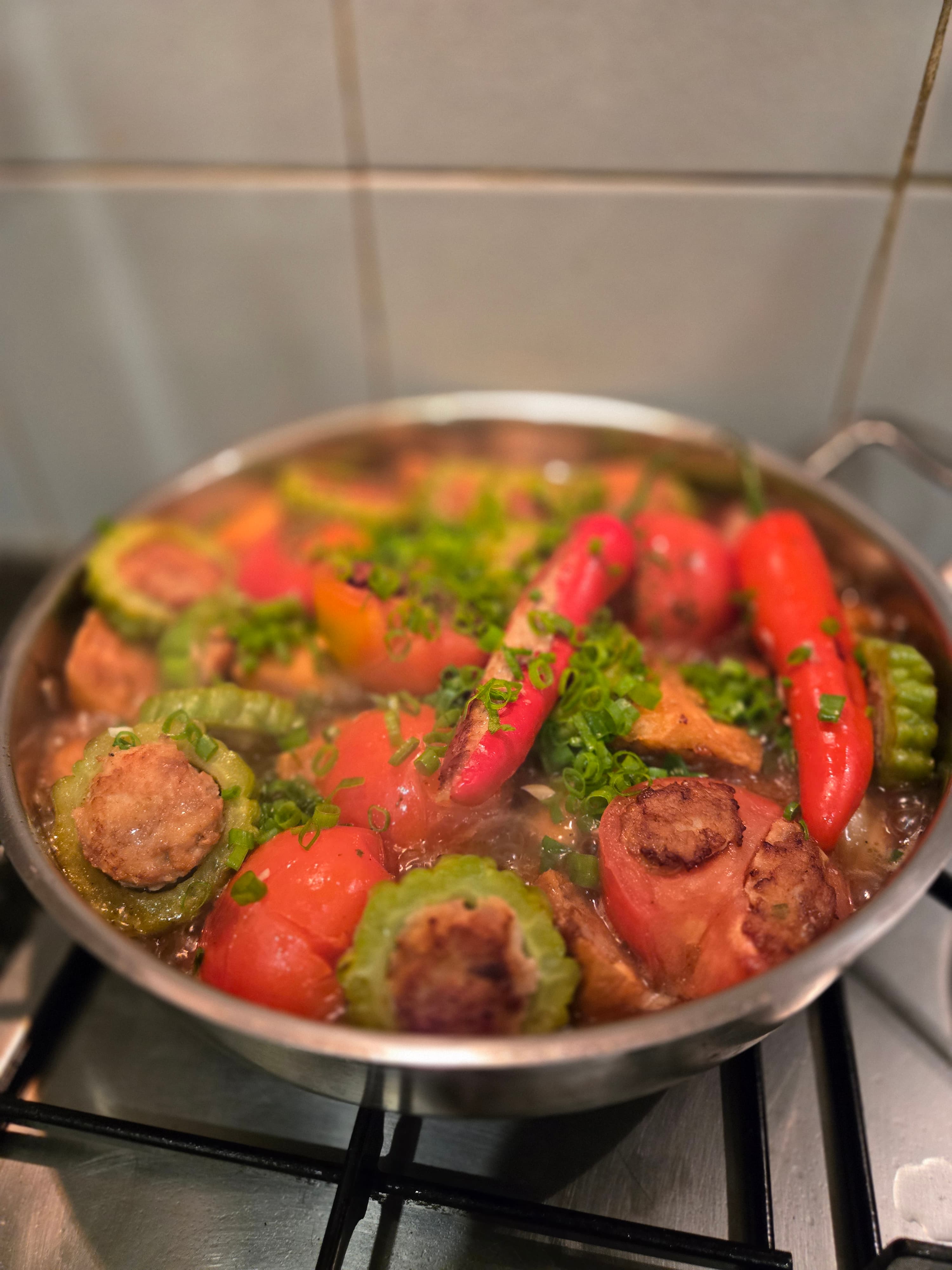 Yong Tau Foo simmering in a stainless pan on a gas hob: stuffed chillies, bitter gourd rings showing the seared pork-and-fish paste, stuffed tomatoes, and scattered spring onion in a clear stock